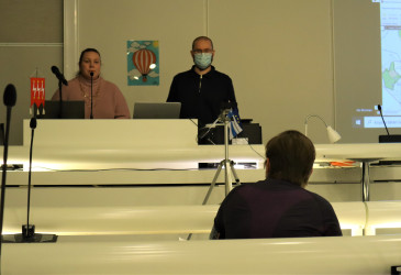 keskustelijoista selin oleva istumassa ja kameraan katsovat seisten valtuustosalin etualalla.