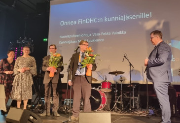 ihmisiä lavalla ja kahdella miehellä kukkapuskat sylissä.