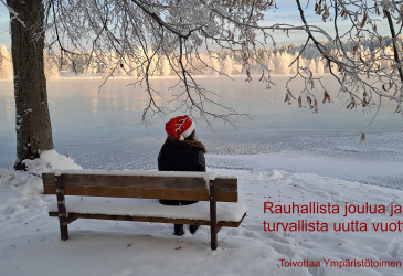 Joulutonttu istuu penkillä selin kuvaajaan veden äärellä rannassa.