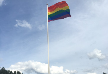 Sateenkaarilippu liehuu Ukonniemen urheilukentän lipputangossa. Taustalla taivas ja muutama pilvi.