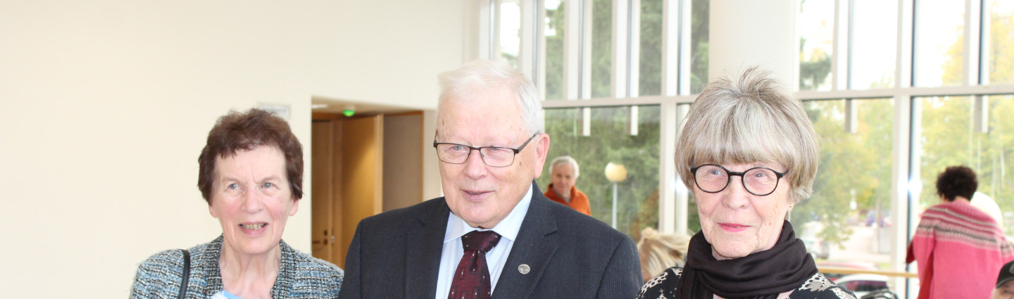 Kolme vanhusta seisomassa, kolmannella kukkapuska kädessä.