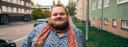 Henkilö kävelee kadulla, hänellä on päällään sininen paita ja punertava huivi. Taustalla näkyy kerrostaloja ja autoja.