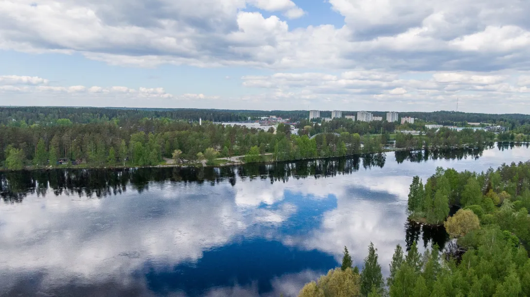 Maisemakuva, jossa etualalla Vuoksen tyyni pinta ja taka-alalla siintävät Mansikkalan tornitalot. 
