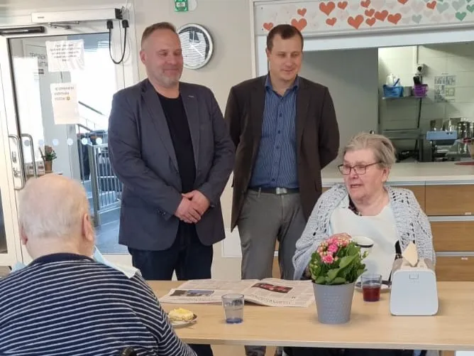 Kaksi miestä seisomassa ja kaksi vanhusta istumassa pöydän ääressä.