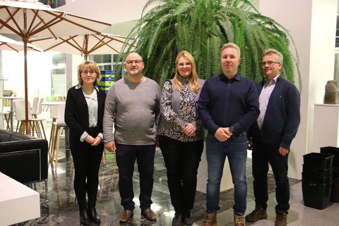 Ryhmäkuva viidestä henkilöstä. He seisovat kaupungintalon aulassa rivissä, heidän takanaan on iso viherkasvi ja tuoleja.